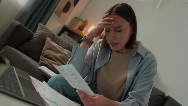 Medium Dutch angle shot of young Caucasian woman sitting on sofa, opening envelope, taking out tax bill or overdue bank invoice, clutching forehead, becoming upset due to financial problems