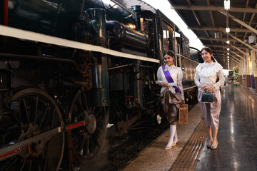 Two attractive women  in traditional Thai dress pose for a photo with a steam train at  the platform of the retro train station