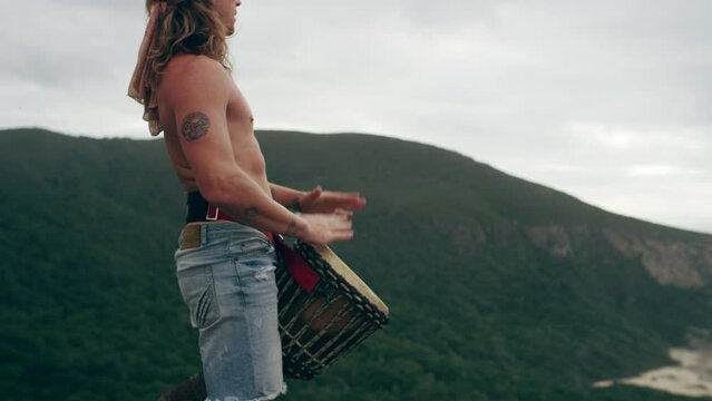Man standing on a mountain by the ocean and playing the Djembe drums