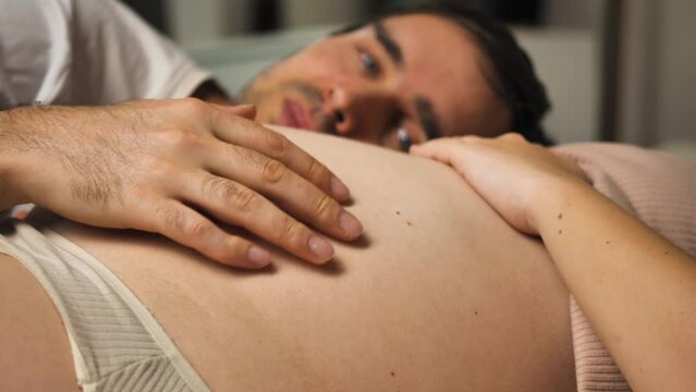 Close-up of the hands of the mother-to-be and her husband, gently stroking the rounded belly. The man lies behind the woman, his head is blurred. A happy family waiting for the birth of a child