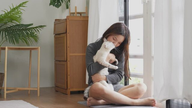 An Asian woman is sitting on a sofa, gently petting her cat, and smiling in her cozy living room