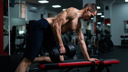 Shirtless man doing dumbbell row to waist on bench at gym. 