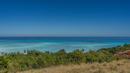 Endless turquoise-aquamarine ocean and clear blue sky. A tiny island in the distance. In the foreground is a meadow with yellowed grass, lush green vegetation. Madagascar. Nosy Iranja.  Copy space.