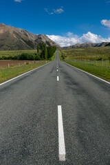 A straight, open road stretches into the distance, flanked by green fields and leading to distant mountains under a bright blue sky.
