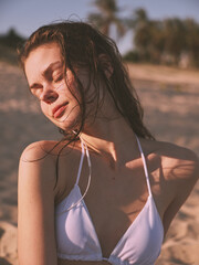 Serene woman in white bikini relaxing on sandy beach with eyes closed and hair blowing in wind