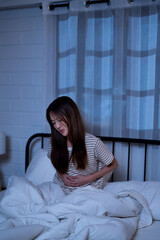 Asian woman in a striped shirt casual pajama sit in bed at night holding her stomach in pain from gastritis or appendicitis reflecting nighttime discomfort after agony awake from symptom