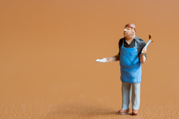 Miniature people , A chef is wearing a blue apron over a black shirt and light-colored pants.  The background is a solid,