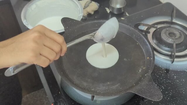 In the kitchen, a woman makes dosa for breakfast by pouring white batter into a black cooking pan and spreads it using a ladle into a perfect round shape in the morning