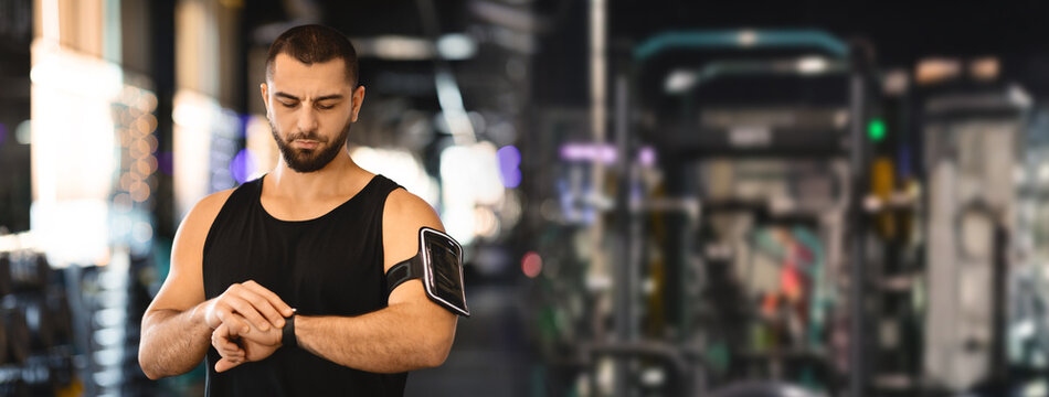 A young man with a beard is wearing a black tank top and checking his smartwatch. He is standing in a gym, with exercise equipment blurred in the background, copy space