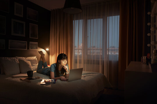Studing at home online. Pretty young woman using laptop while lying on the bed at night time.