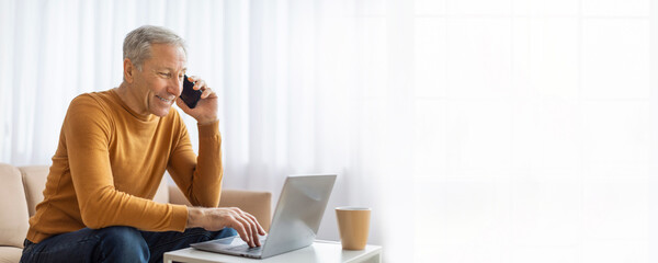 A mature man in a yellow sweater sits on a couch in a bright room while working on a laptop. He is talking on a cell phone and looks at the screen, copy space