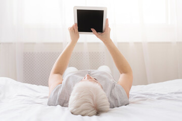 An individual reclining on a bed, engrossed in a tablet screen. The elderly woman appears relaxed...