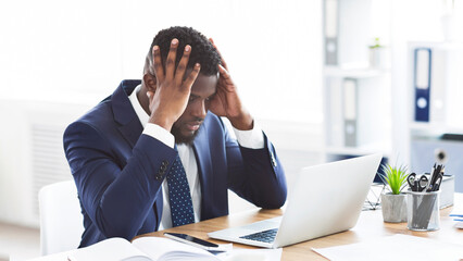 Stressed and overworked african-american young businessman sitting in office with hand on forehead,...