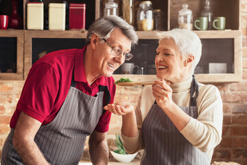 A senior couple stands together in a home kitchen preparing a meal. The man is wearing a red shirt and gray striped apron while chopping vegetables on a cutting board.