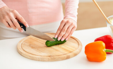 Lady uses a knife to slice a cucumber on a wooden cutting board in a brightly lit kitchen. Nearby on the counter, red and yellow bell peppers are also visible. - Powered by Adobe