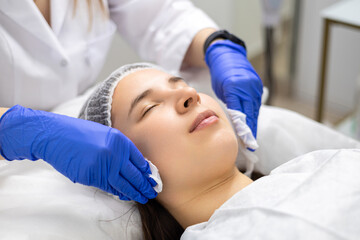 Naklejka premium Close-up shot of a young beautiful woman relaxing in a beauty saloon or spa with eyes closed while the beautician cleanses her face with tissues, to prepare the skin for a beauty treatment.