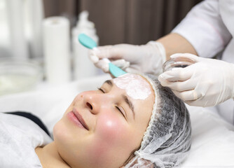 Close-up side profile portrait of a young woman relaxing in a beauty salon while a dermatologist applies a clay face mask on her face.