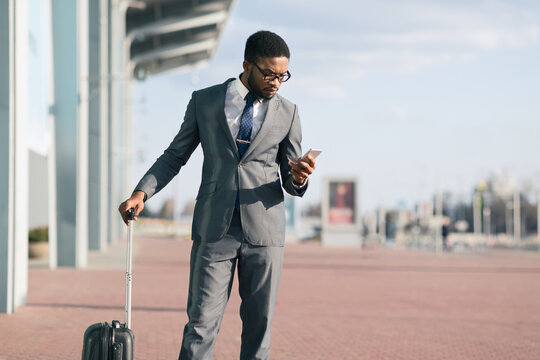 African American professional man with a suitcase is using his phone outside an airport. The scene is dynamic and modern, reflecting business travel and the fast-paced corporate world.