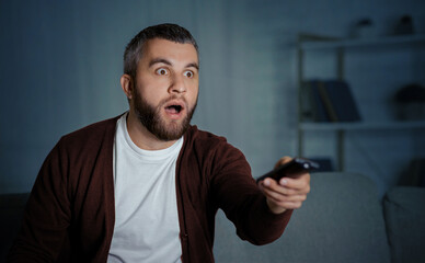 A man with a shocked expression is sitting on his couch at home in the evening, holding a TV remote...