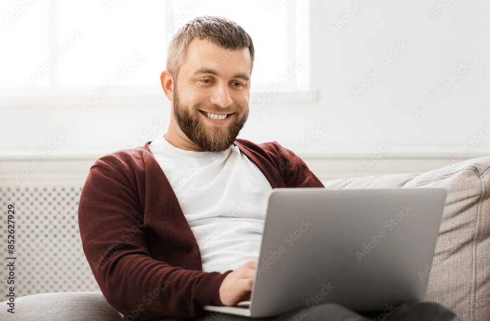 Poster a man is seated on a comfortable couch, focused on a laptop screen in front of him. he appears engag