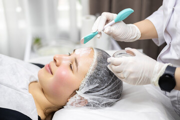 Close-up side profile portrait of a young woman enjoying a beauty treatment. The dermatologist applies a clay face mask on her face with a spatula.