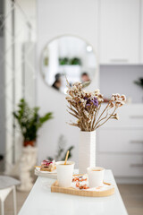 A bright and modern beauty salon interior featuring a white table with a vase of dried flowers, two cups of coffee, and a some delicious treats..