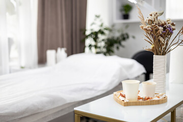 Modern, minimalist beauty salon interior, a white table displays a vase of dried flowers, three cups of coffee or tea and some sweets. On background can be seen a procedure table.
