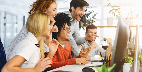 A team of cheerful multiethnic co-workers is gathered around a computer screen, exuding excitement and jubilation. Their expressions of joy and elation, along with positive gestures