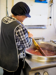 Woman stirring a large pot in commercial kitchen, wearing checkered shirt and black apron.