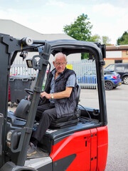 Elderly man operating a red forklift truck in an industrial yard on a sunny day, focused and determined.