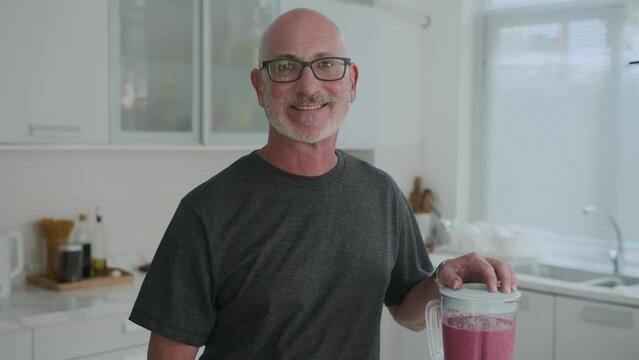 Tilt up portrait shot of mature Caucasian man in glasses making healthy smoothie from fruits and looking at camera at kitchen