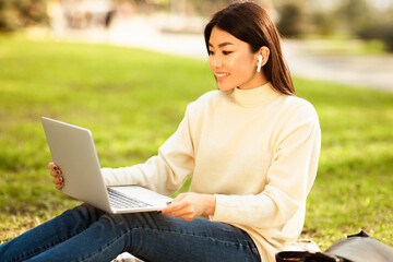 Asian woman is seated on green grass, focusing on her laptop computer and earbuds. She is typing on...