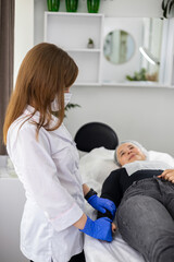 Fototapeta premium Beautiful young woman lying down on a procedure table and smiling while looking at the beautician which stays right near her, after a cosmetic procedure. 
