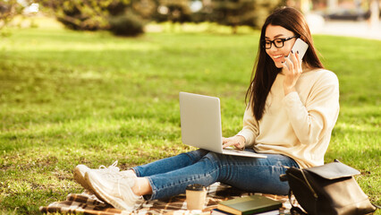A young Asian woman is sitting on a plaid blanket in a park on a sunny afternoon, multitasking by...