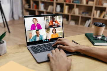 Guy uses a laptop to participate in a video call with four other people. The call is taking place in a home office setting, with a bookshelf in the background.