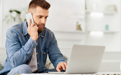 A man seated on the floor, multitasking by using a laptop and talking on a cell phone.