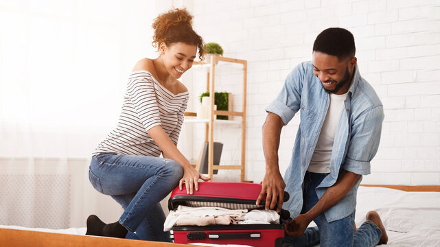 African American man and a woman are shown in a bedroom, unpacking luggage on top of a bed. They are sorting clothes and personal items from their suitcases.