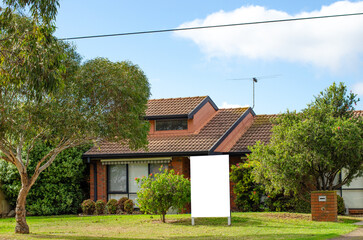Background texture of a mockup real estate sign at the front yard garden of a single-story suburban house with a gabled roof. Empty blank white signage of a residential property in Australia