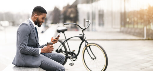African American businessman is seated near his bike, using his smartphone. The outdoor environment is serene yet professional, highlighting a balance between work and relaxation.