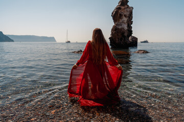 A woman in a red dress stands on a beach with a rocky shoreline in the background. The scene is serene and peaceful, with the woman's red dress contrasting against the natural elements of the beach.