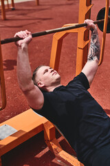 Caucasian male athlete trains arms and chest with barbell, outdoor workout in summer park