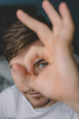 Obraz premium Portrait of a man. He covers his face with his hand. Shows the OK sign. He looks into his folded fingers like into a peephole. The hand is blurred in the foreground