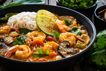 Tom Yam Kung soup - Thai soup with shrimps and rice noodles served in black bowl with chopsticks on wooden table
