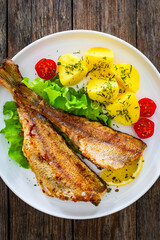 Fried sea bass served on lettuce with boiled potatoes and  lemon on white plate on wooden table
