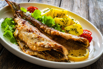 Fried sea bass served on lettuce with boiled potatoes and  lemon on white plate on wooden table
