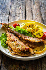 Fried sea bass served on lettuce with boiled potatoes and  lemon on white plate on wooden table
