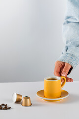 Hand reaching for a yellow coffee cup with golden coffee capsules and beans on a white background