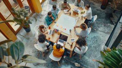 A diverse group of people are gathered around a large wooden table in a modern office setting. They are collaborating on a project, using laptops, a whiteboard, and other materials. The team appears t
