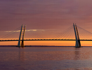 Obraz premium A breathtaking view of a cable-stayed bridge during a stunning sunset, with vibrant colors reflecting on calm waters. The dramatic sky adds to the beauty and serenity of the scene.