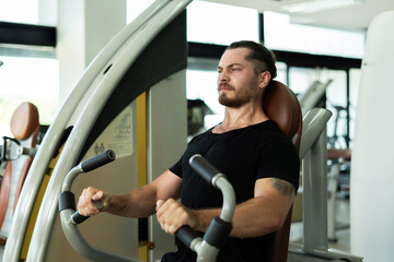 A man is sitting on a machine in a gym. He is wearing a black shirt and has a beard